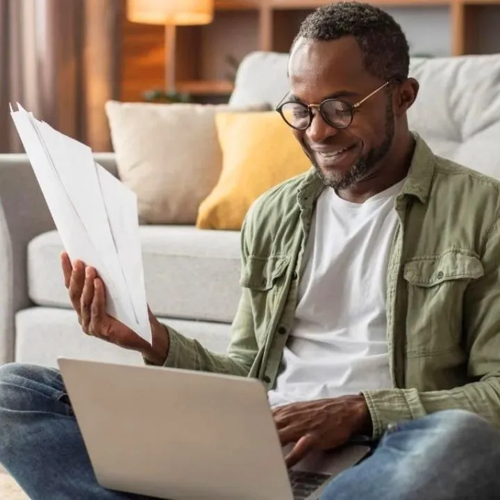 Propriétaire souriant qui consulte ses documents sur un canapé avec son ordinateur portable